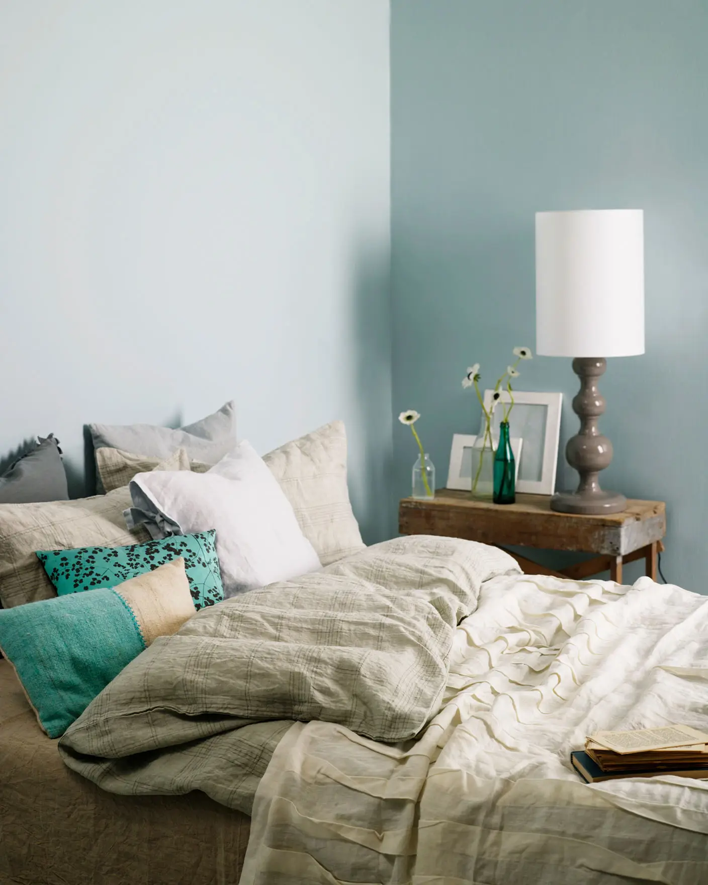 Inviting bedroom with layered neutral bedding, turquoise accent pillows, and a rustic wooden side table, set against muted, greenish blue wall painted with Jotun 5225 Lake View.