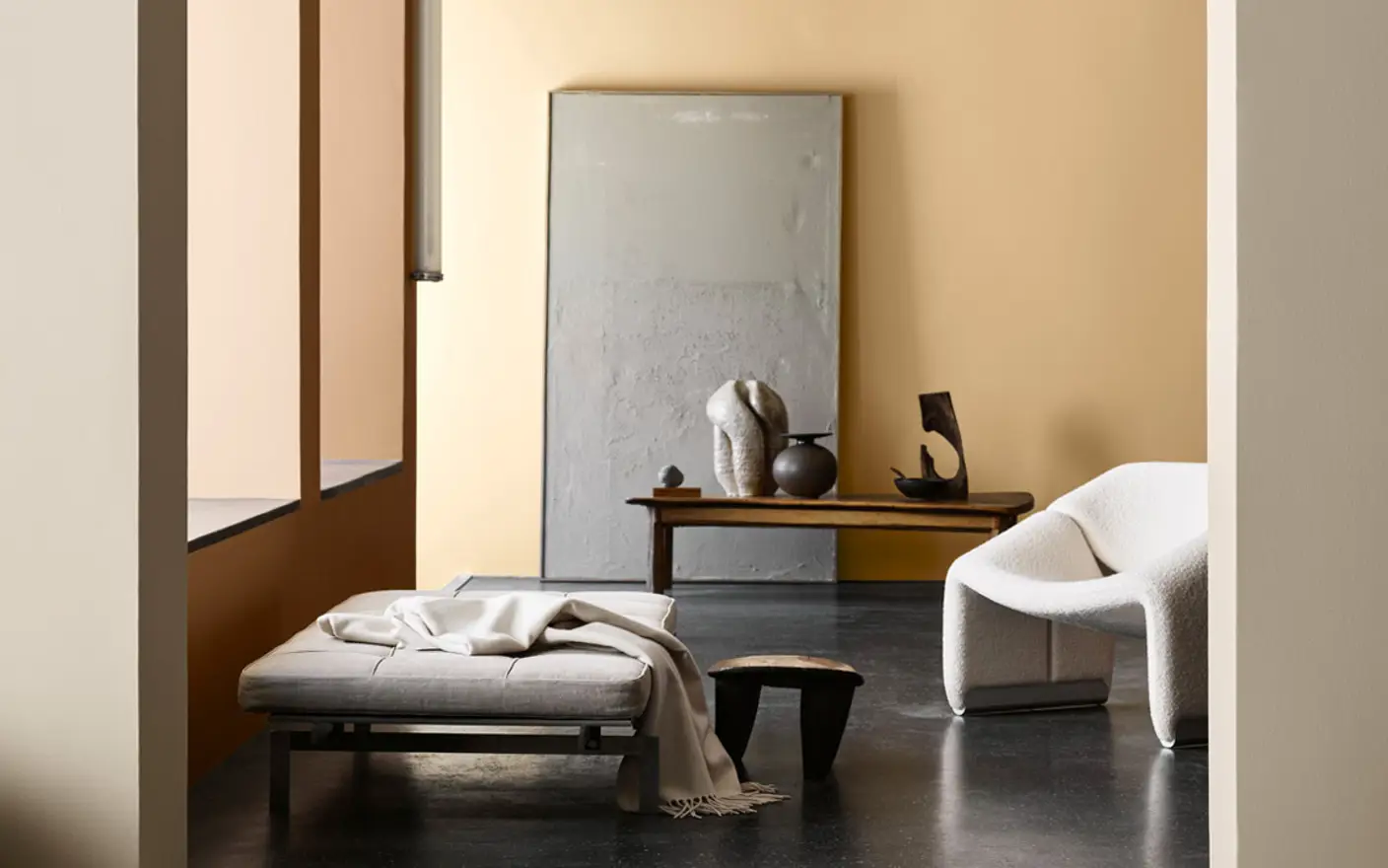 Moderne stue med minimalistisk design, inkludert en beige stol, grå sofa med pledd, trebenk med skulpturer, og en stor speil. Veggene er malte i lyse toner for en varm atmosfære