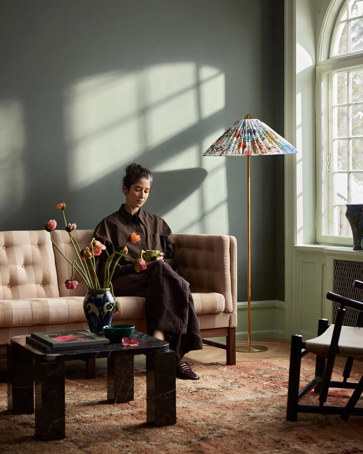 Woman sitting in a sofa with sunlight streaming through a window. The background wall is painted with the dark, muted green colour 7613 Northern Mystic from Jotun.